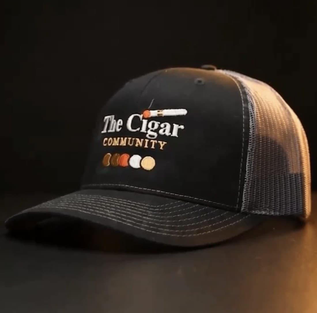 The Cigar Community trucker cap in black, with the embroidered lockup across the front panel and a pale mesh back