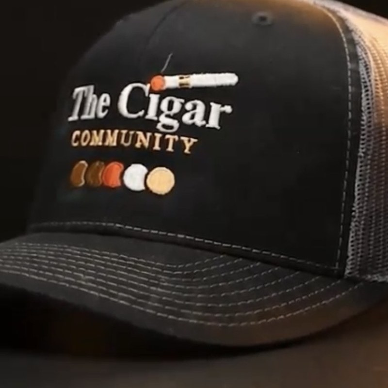Close-up of the embroidered Cigar Community lockup on the black front panel — the wordmark, a lit cigar, and the shade row beneath it